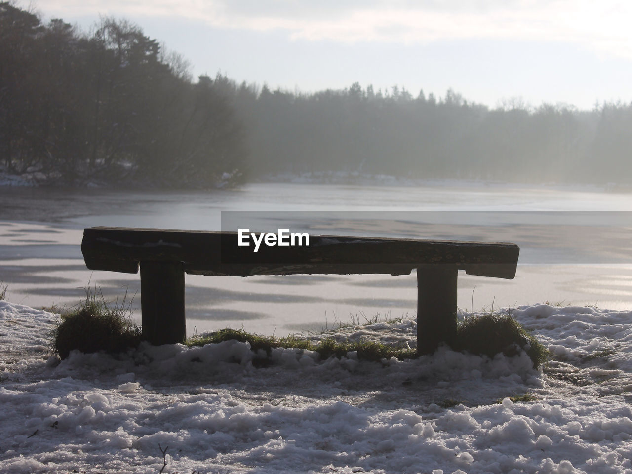 Empty bench on snow covered field against lake and trees