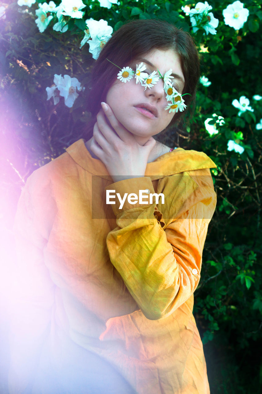 Woman with daisies on eyes while touching neck against plants