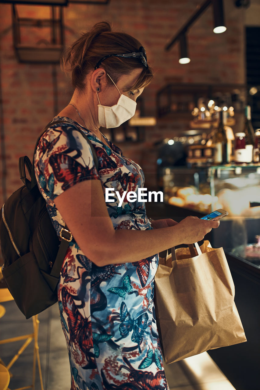 Mature woman wearing mask using mobile phone standing in cafe