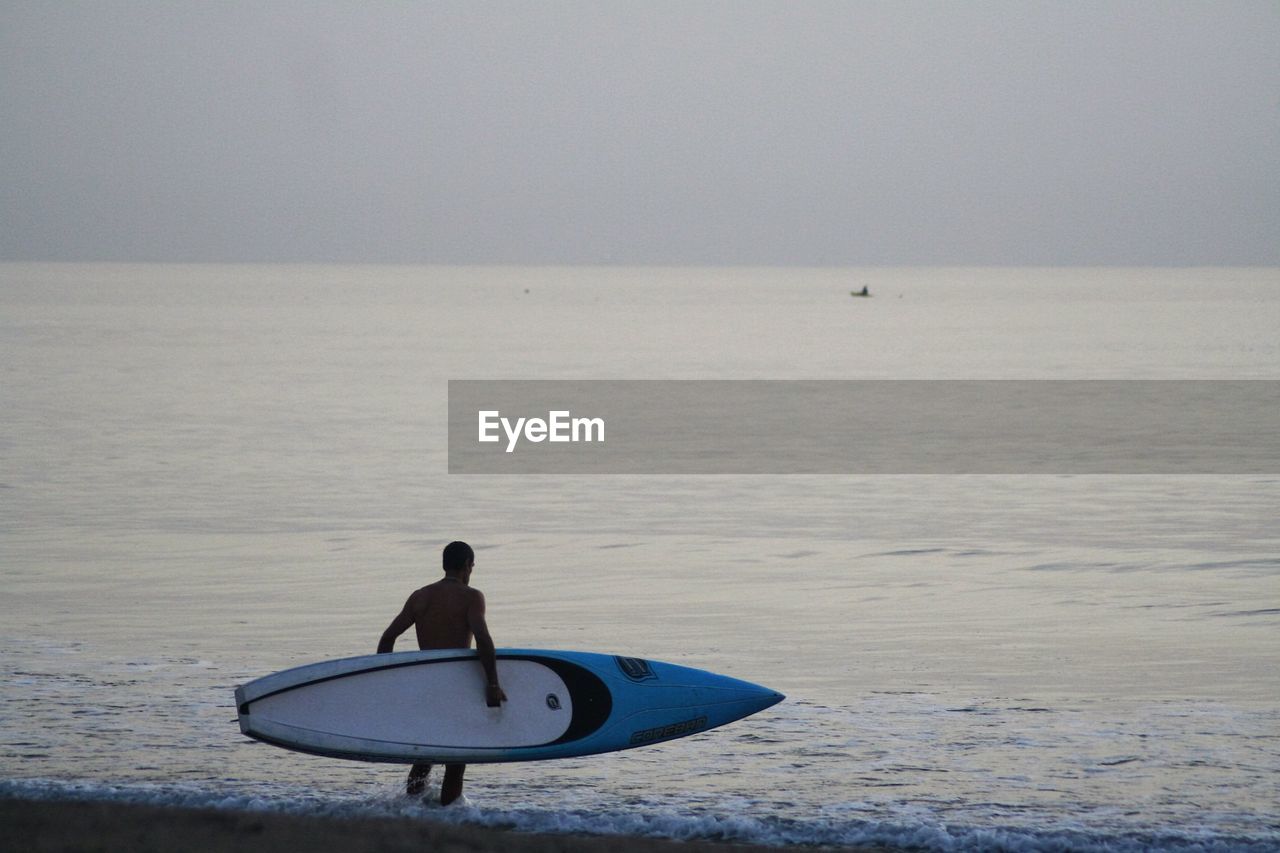Rear view of man carrying surfboard on shore against sky