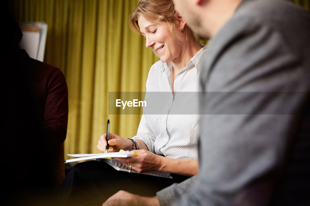 Smiling entrepreneur writing in book while sitting by male colleague during office seminar