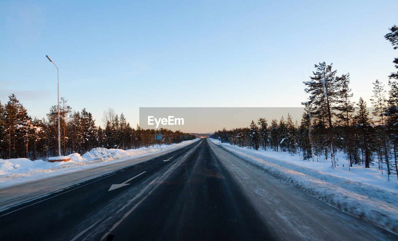 SNOW COVERED ROAD AMIDST BARE TREES AGAINST SKY