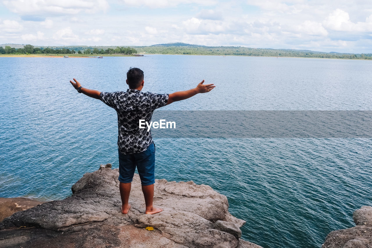 Rear view of man with arms outstretched standing on rock by sea against sky