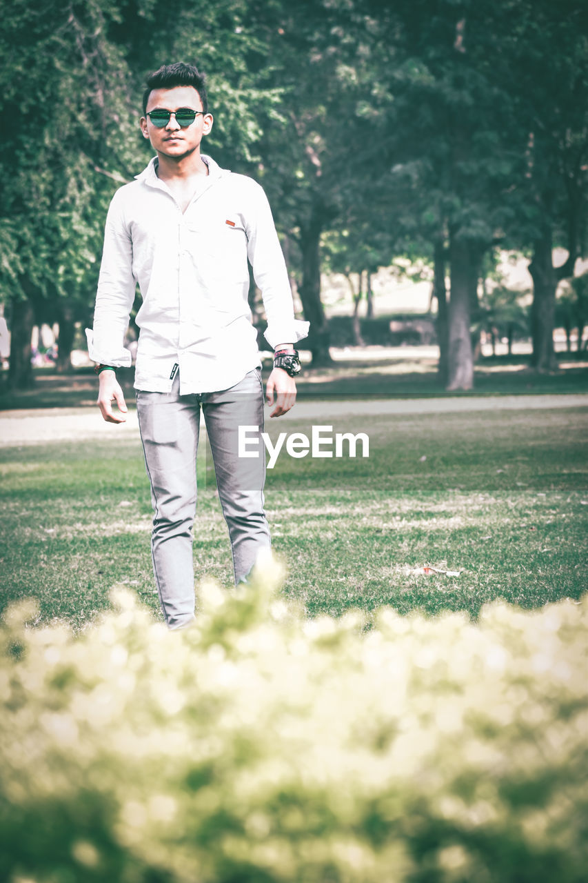one person, plant, sunglasses, spring, young adult, nature, adult, glasses, grass, fashion, men, white, casual clothing, green, lifestyles, front view, tree, full length, leisure activity, portrait, day, sunlight, standing, park, outdoors, park - man made space, sports, emotion, clothing, happiness, smiling