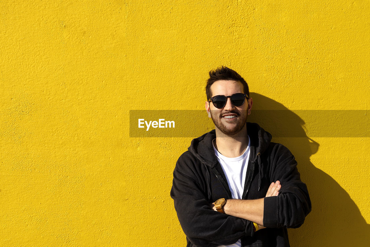 Young bearded man standing against a yellow wall with crossed arms