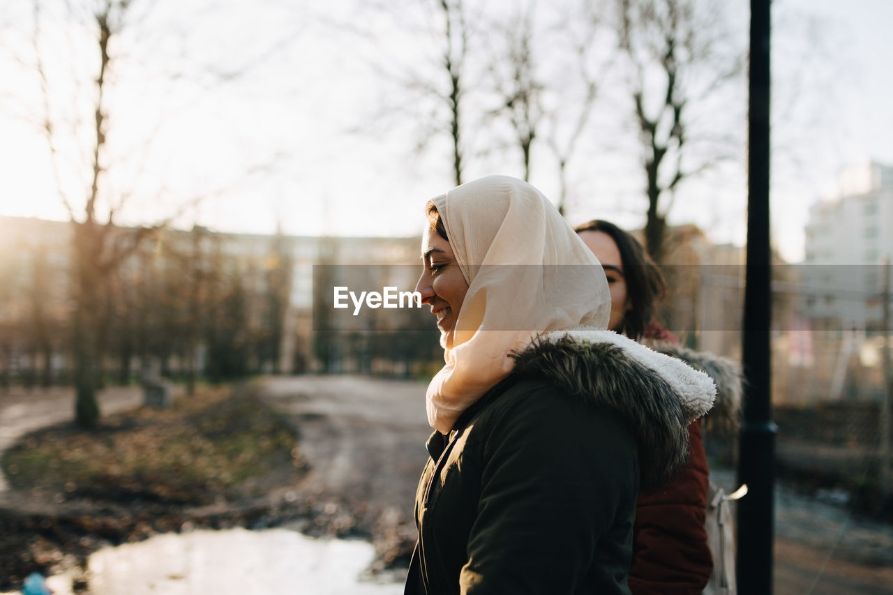 Side view of smiling young muslim woman with female friend walking in city during sunset