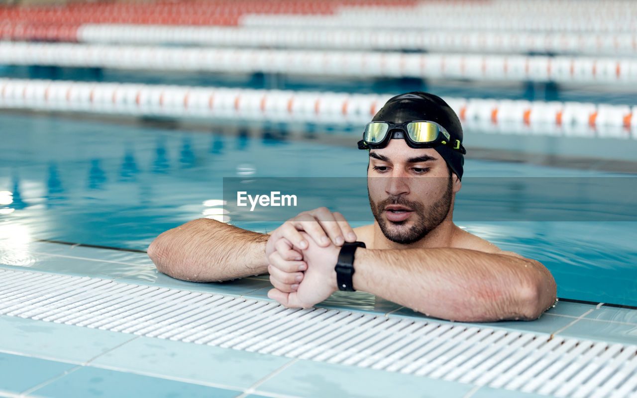Bearded male athlete in swimming cap and goggles watching time on wristwatch while leaning on poolside