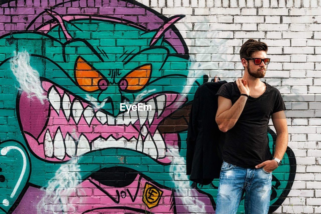 Handsome young man wearing sunglasses while standing against graffiti wall