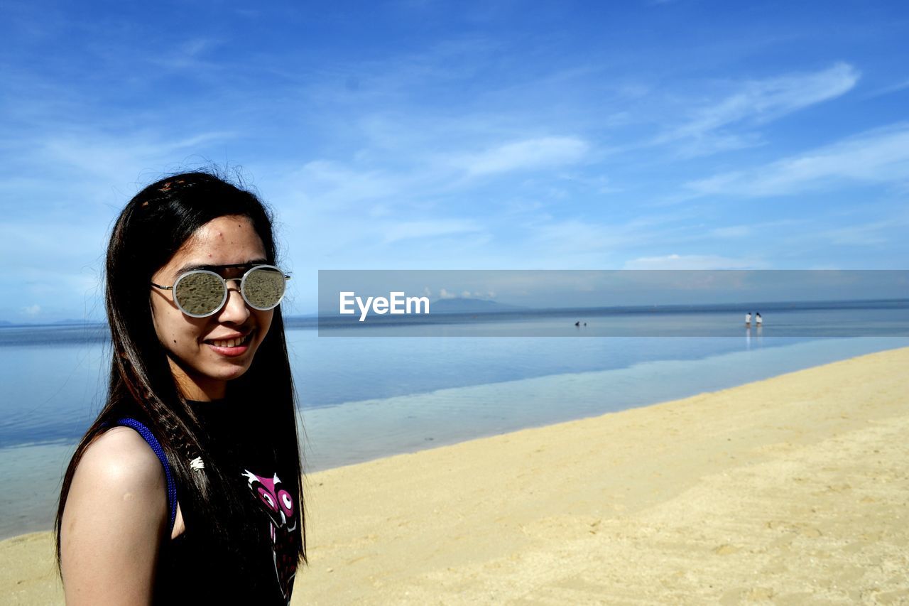 PORTRAIT OF SMILING WOMAN AT BEACH AGAINST SKY