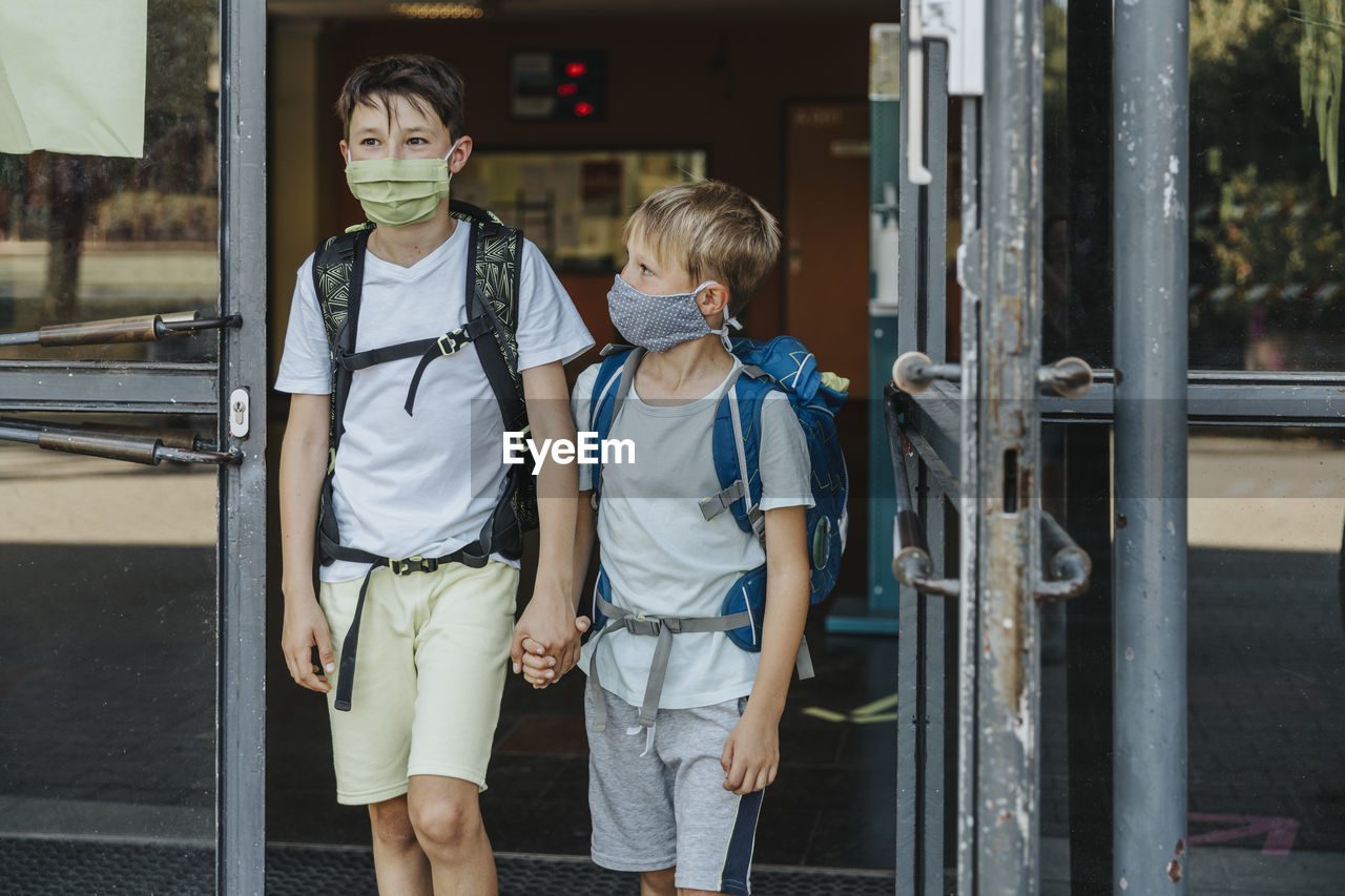 Brothers wearing protective face mask holding hands coming out of school building