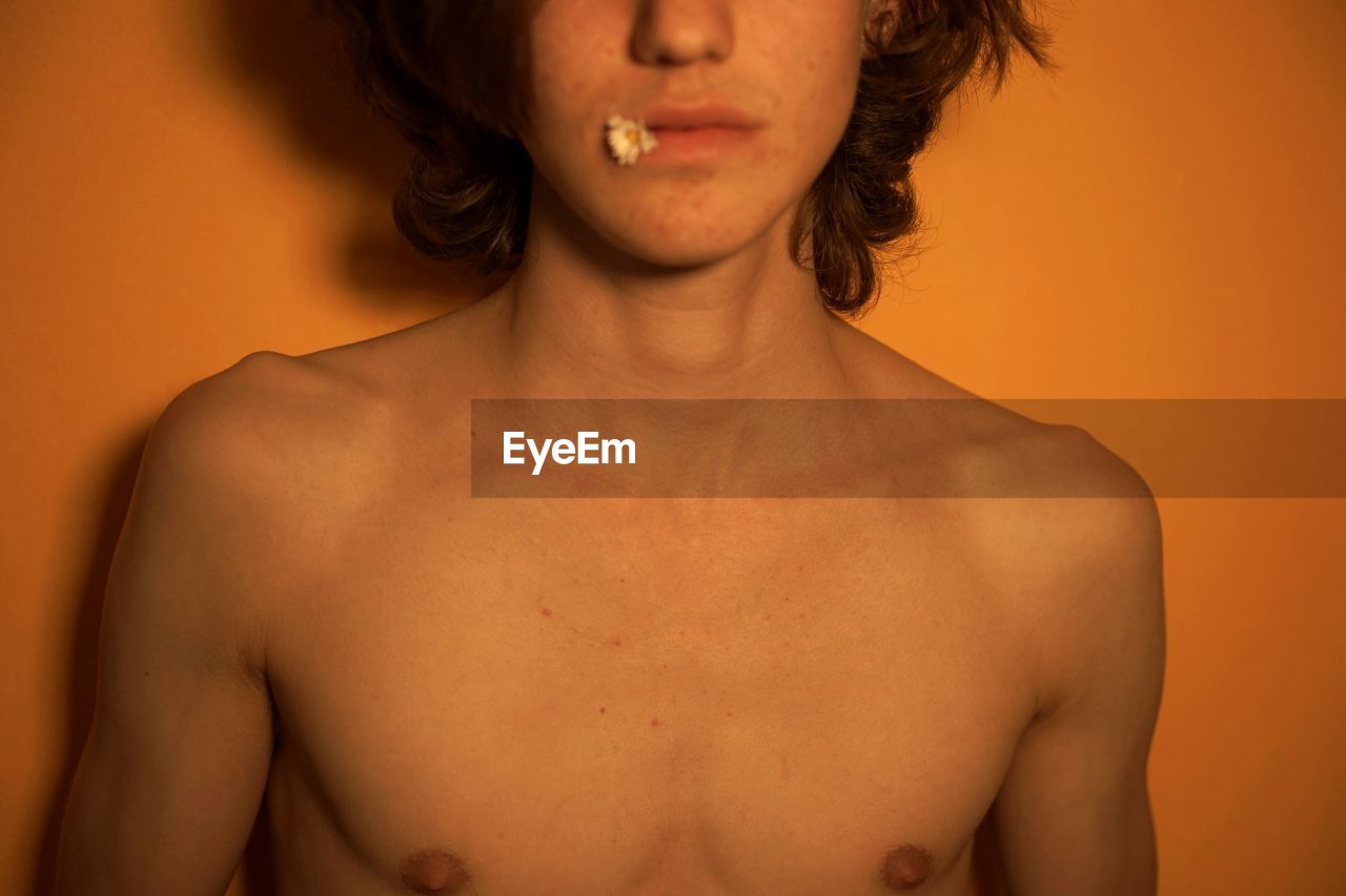 Midsection of shirtless teenage boy standing by orange wall