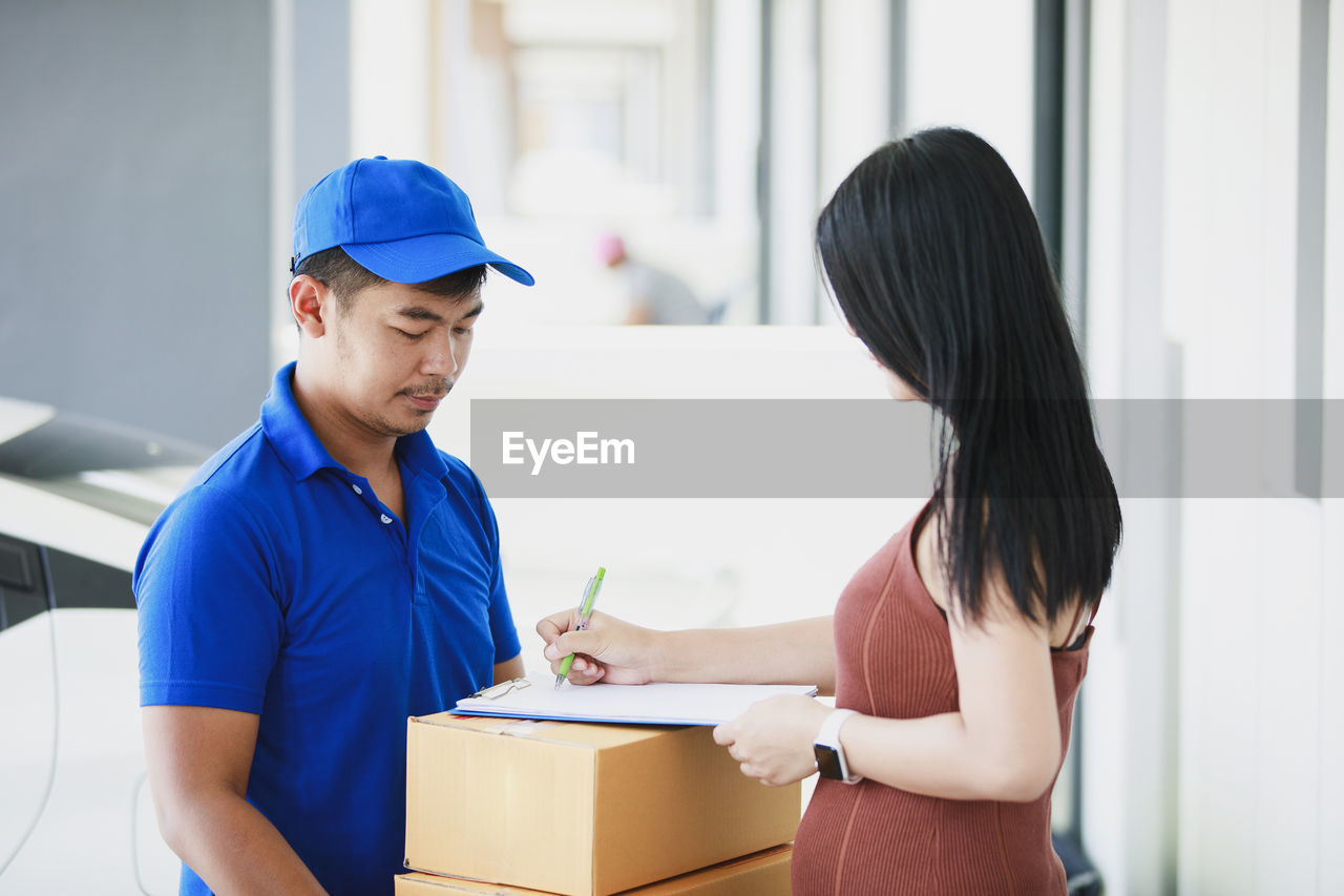 Blue delivery man holding a cardboard box while beautiful young woman putting signature in clipboard