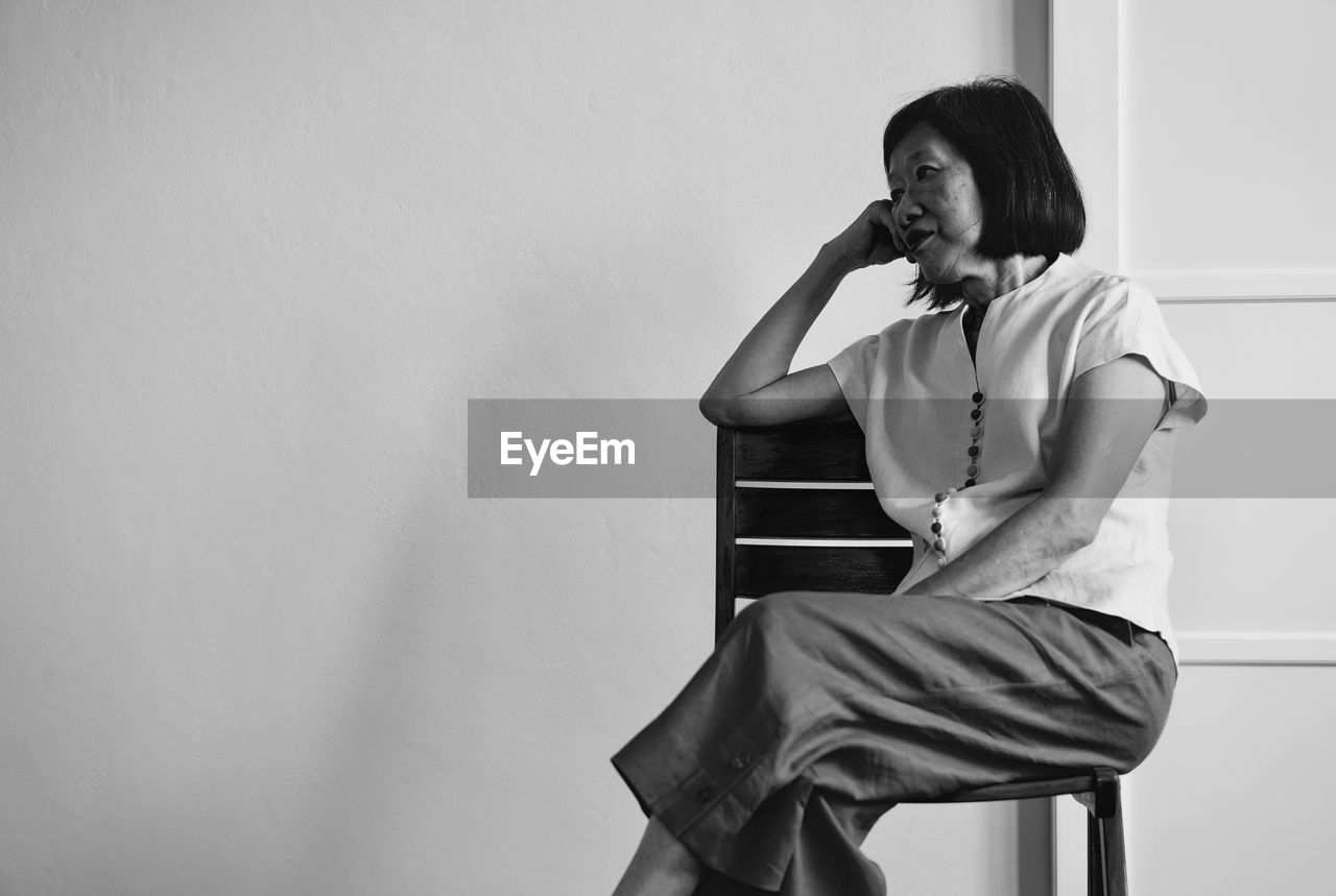 Senior woman looking away while sitting on chair against wall