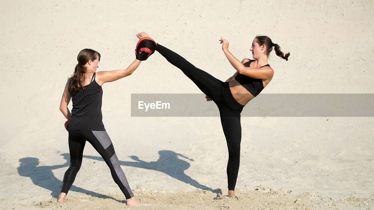 Young women in black fitness suits are engaged in a pair, work out kicks, on a deserted beach