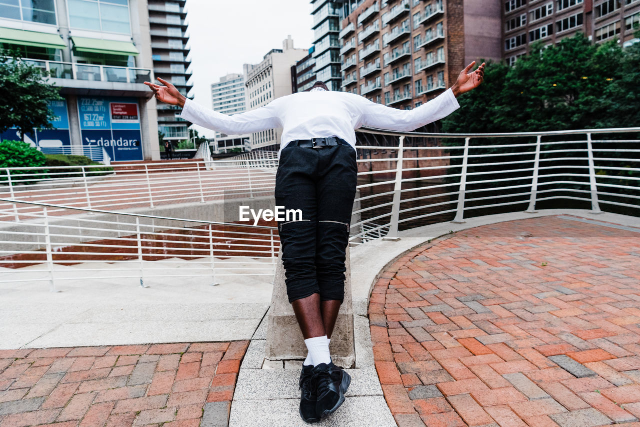 REAR VIEW OF MAN STANDING ON FOOTPATH BY STREET AGAINST BUILDINGS