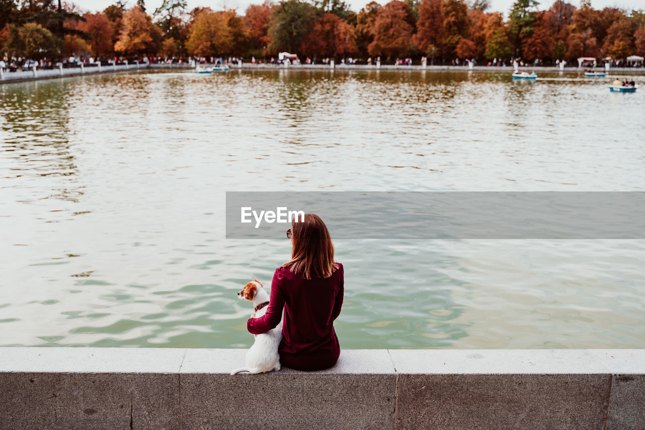 Rear view of woman with dog sitting by lake against trees