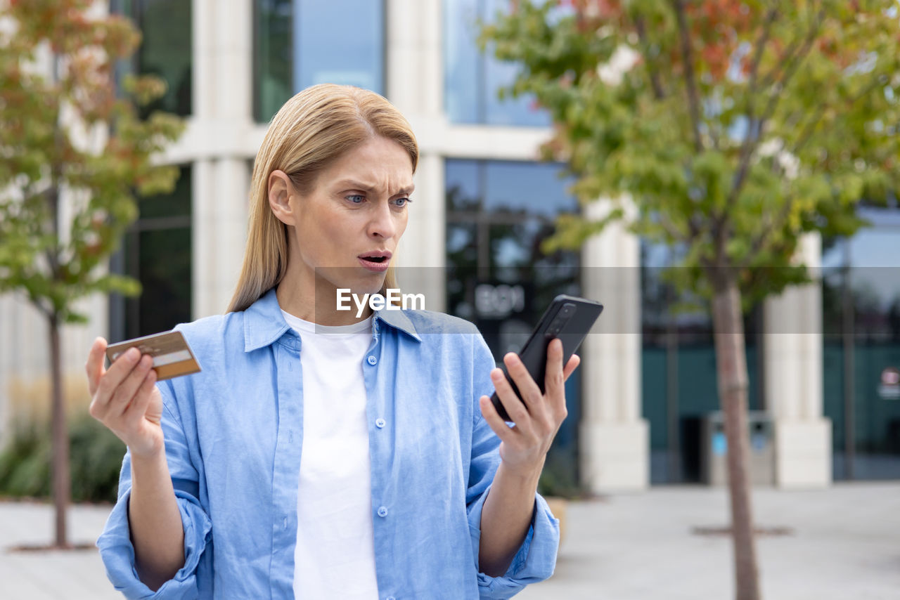 technology, wireless technology, communication, smartphone, one person, mobile phone, portable information device, adult, city, women, telephone, architecture, young adult, spring, using phone, street, looking, clothing, front view, blond hair, city life, holding, portability, day, standing, person, waist up, business, outdoors, lifestyles, casual clothing, building exterior, outerwear, business finance and industry, activity, internet, built structure, female, city street, portrait, focus on foreground, computer network, jacket, convenience, shirt, businesswoman, hairstyle, answering, reading, mobile app
