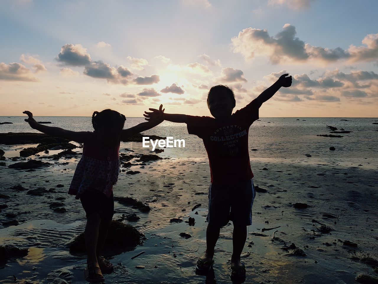 Silhouette siblings with arms outstretched standing at beach against sky