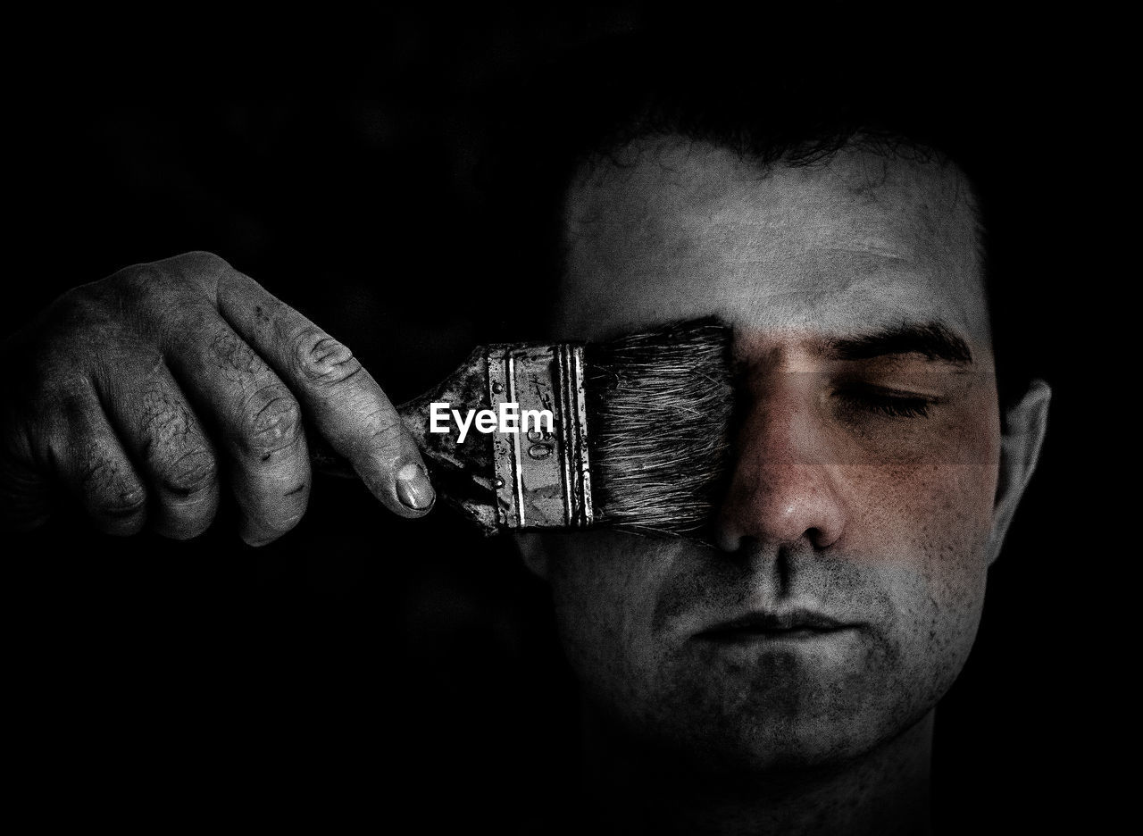 Young man holding paint brush over closed eyes against black background