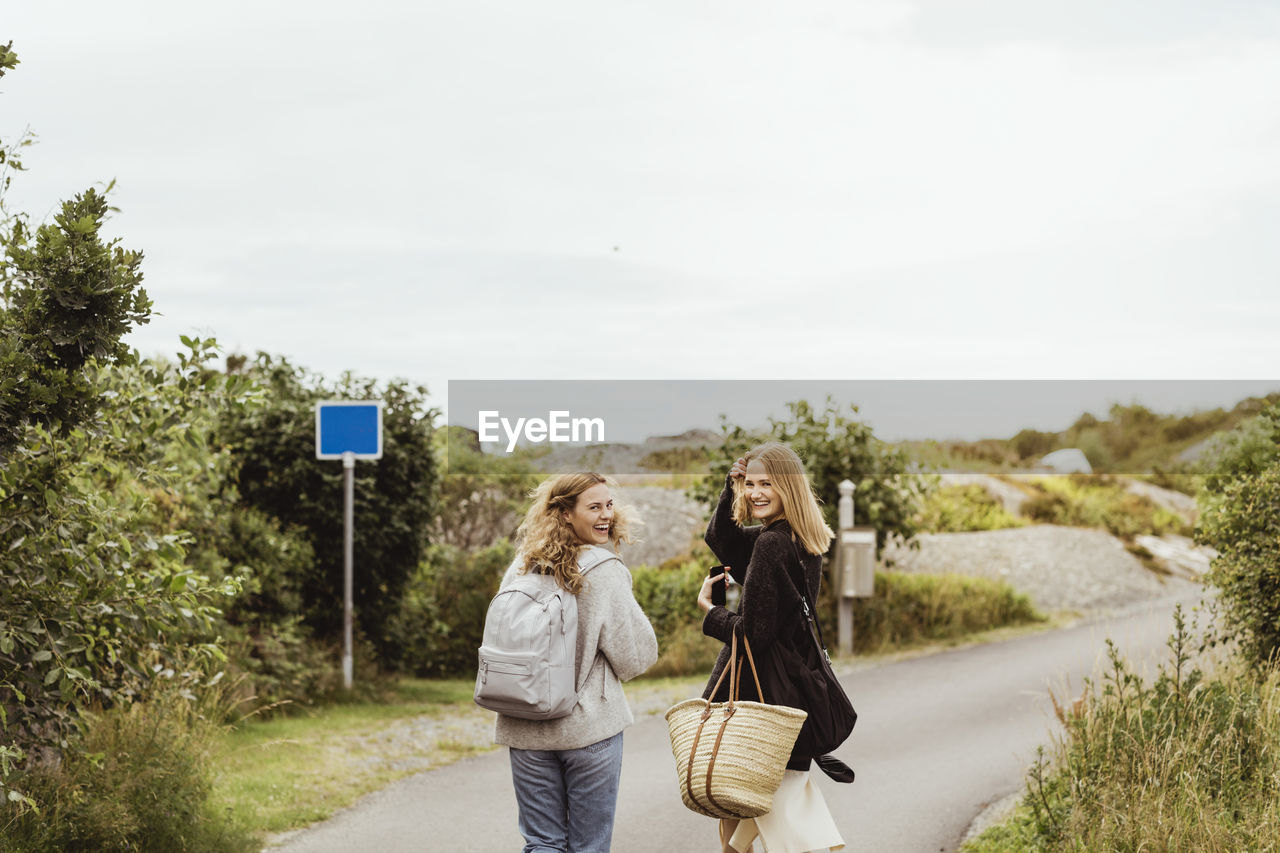 Smiling female friends looking over shoulder while walking on road during summer vacation