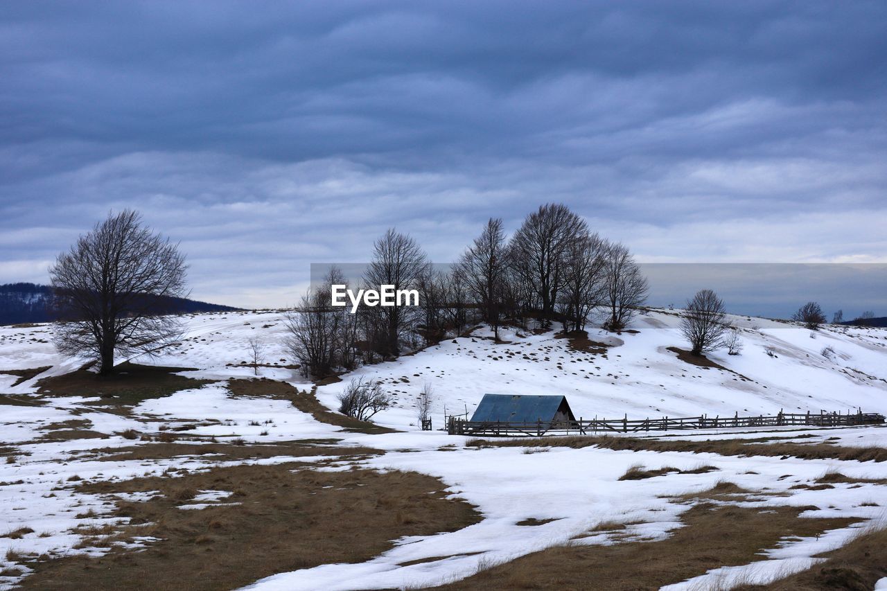 Old wooden house in the distance on a field covered in snow and dark storm clouds in the sky