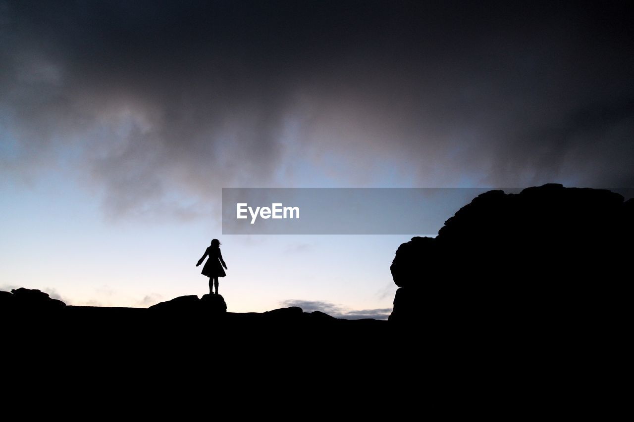Silhouette woman standing on rocks against cloudy sky at sunset