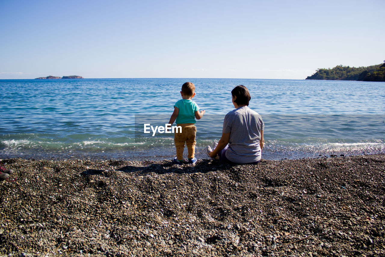 REAR VIEW OF BOYS SITTING ON BEACH AGAINST SKY