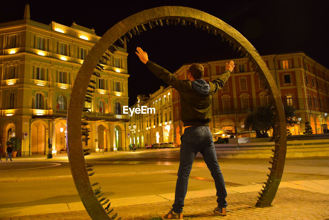 Rear view of man standing on footpath by built structure at night