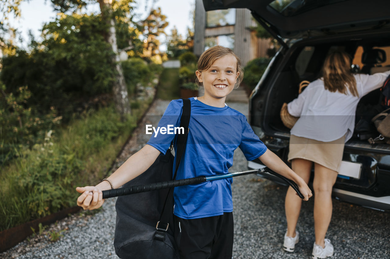 Portrait of smiling boy holding ice hockey stick with mother loading car trunk