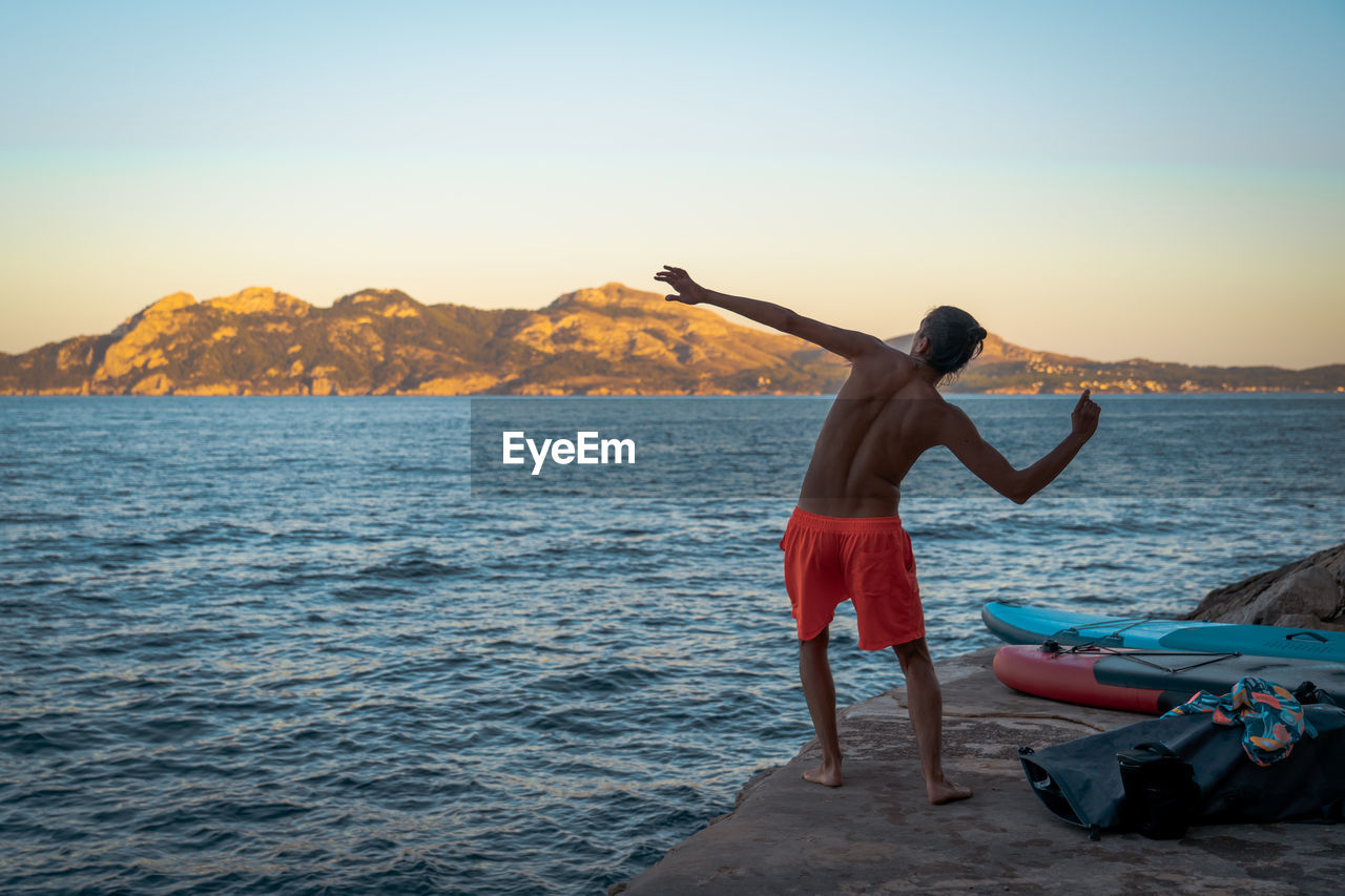 Rear view of shirtless man throwing rock to sea