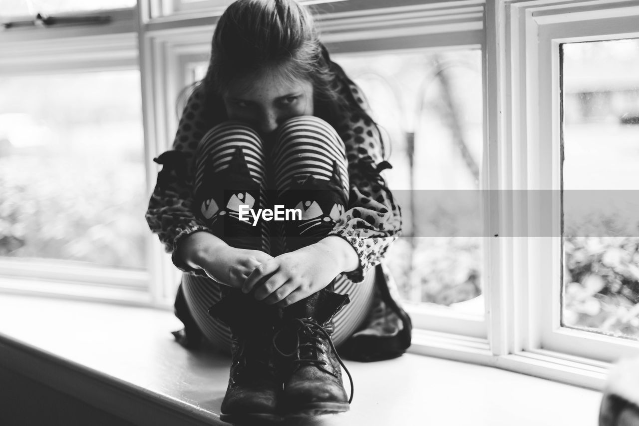 Close-up of girl hugging knees while sitting on window sill