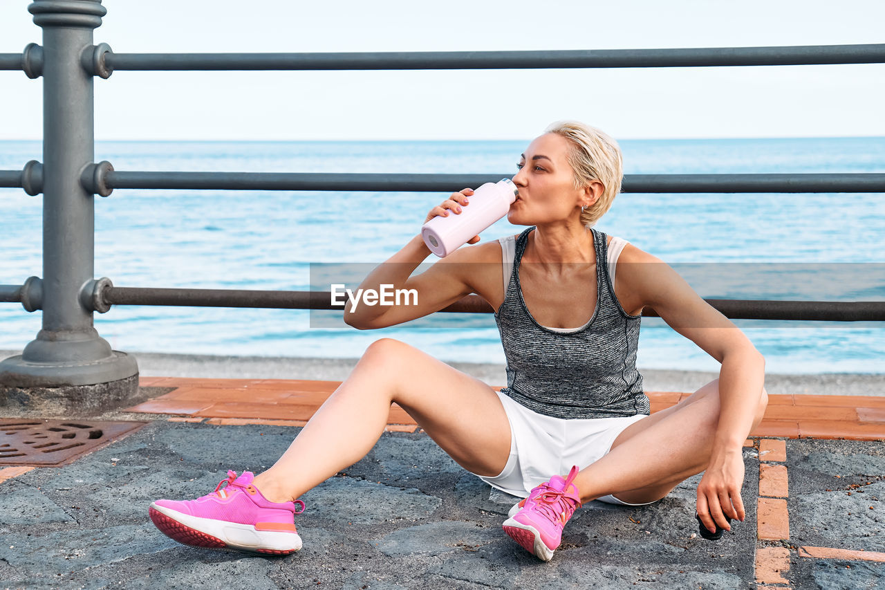 Fit woman relaxes and drinking water on seaside promenade after running and training.