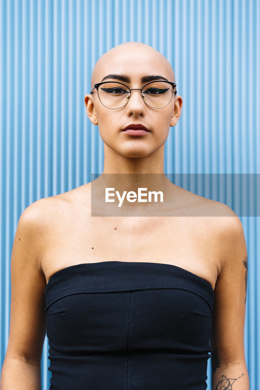 Confident young woman with shaved head wearing eyeglasses in front of blue wall