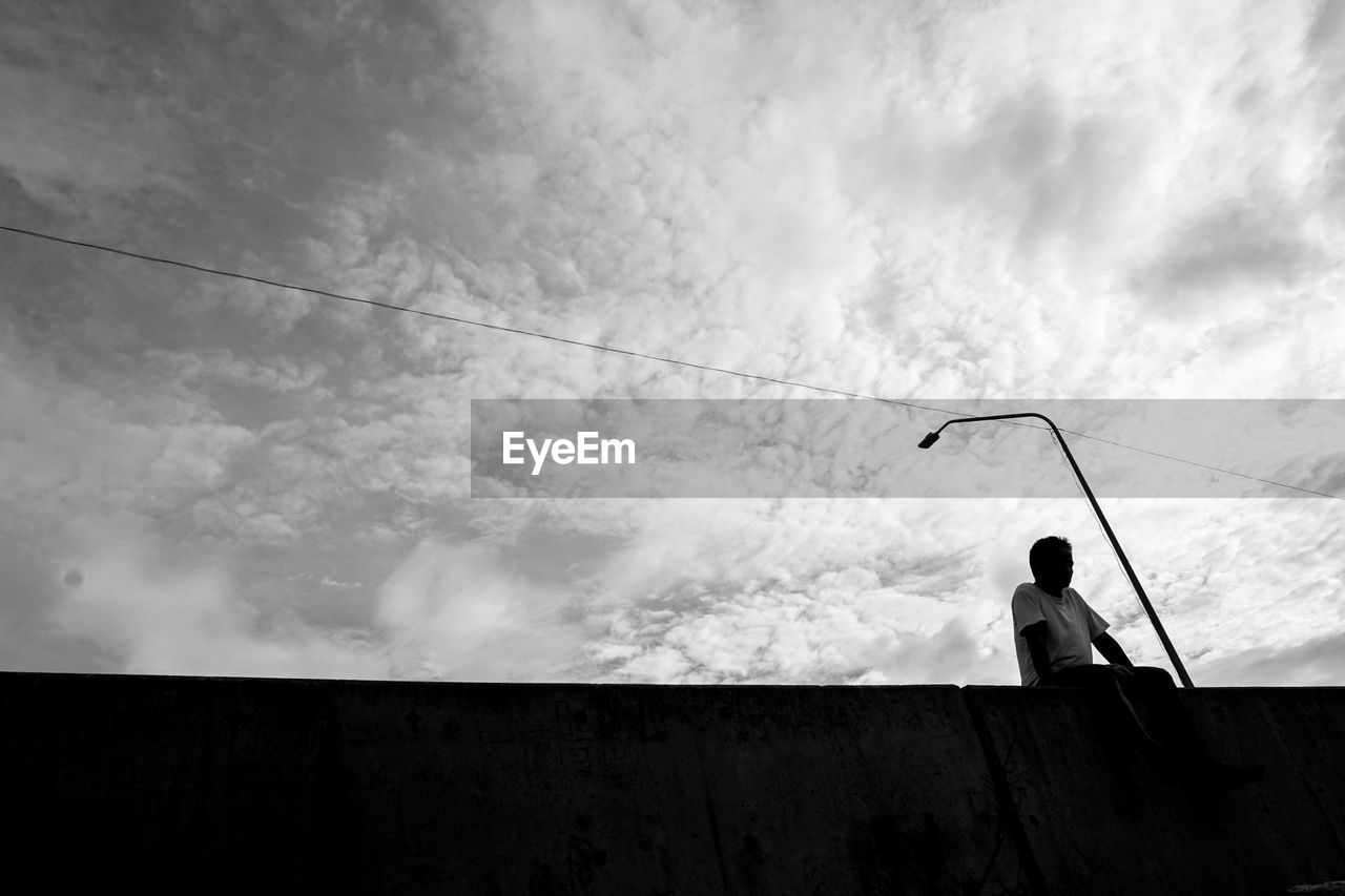 Low angle view of man sitting on railing against cloudy sky