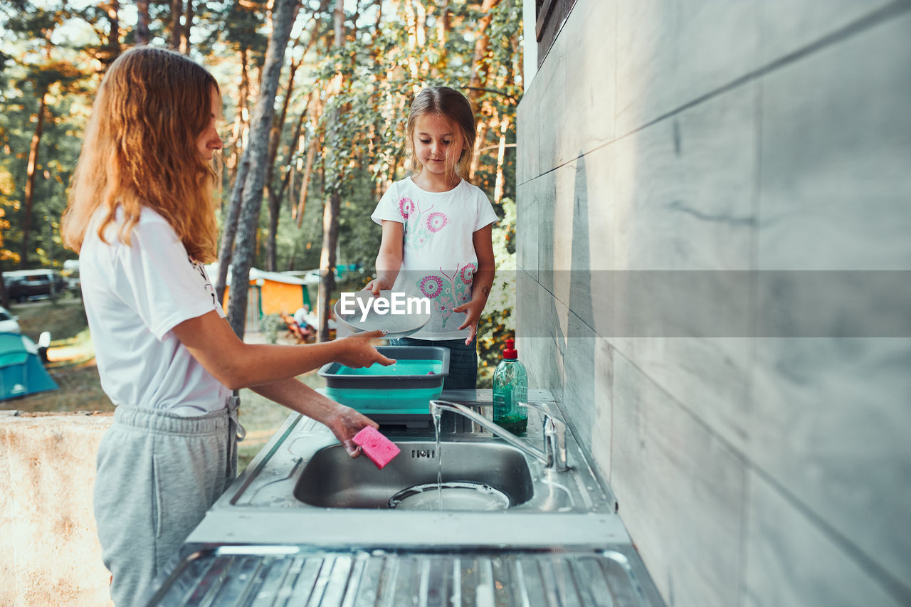 Teenager girl washing up the dishes pots and plates with help her younger sister