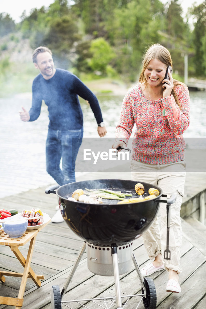 Happy woman using mobile phone while barbecuing with man on pier