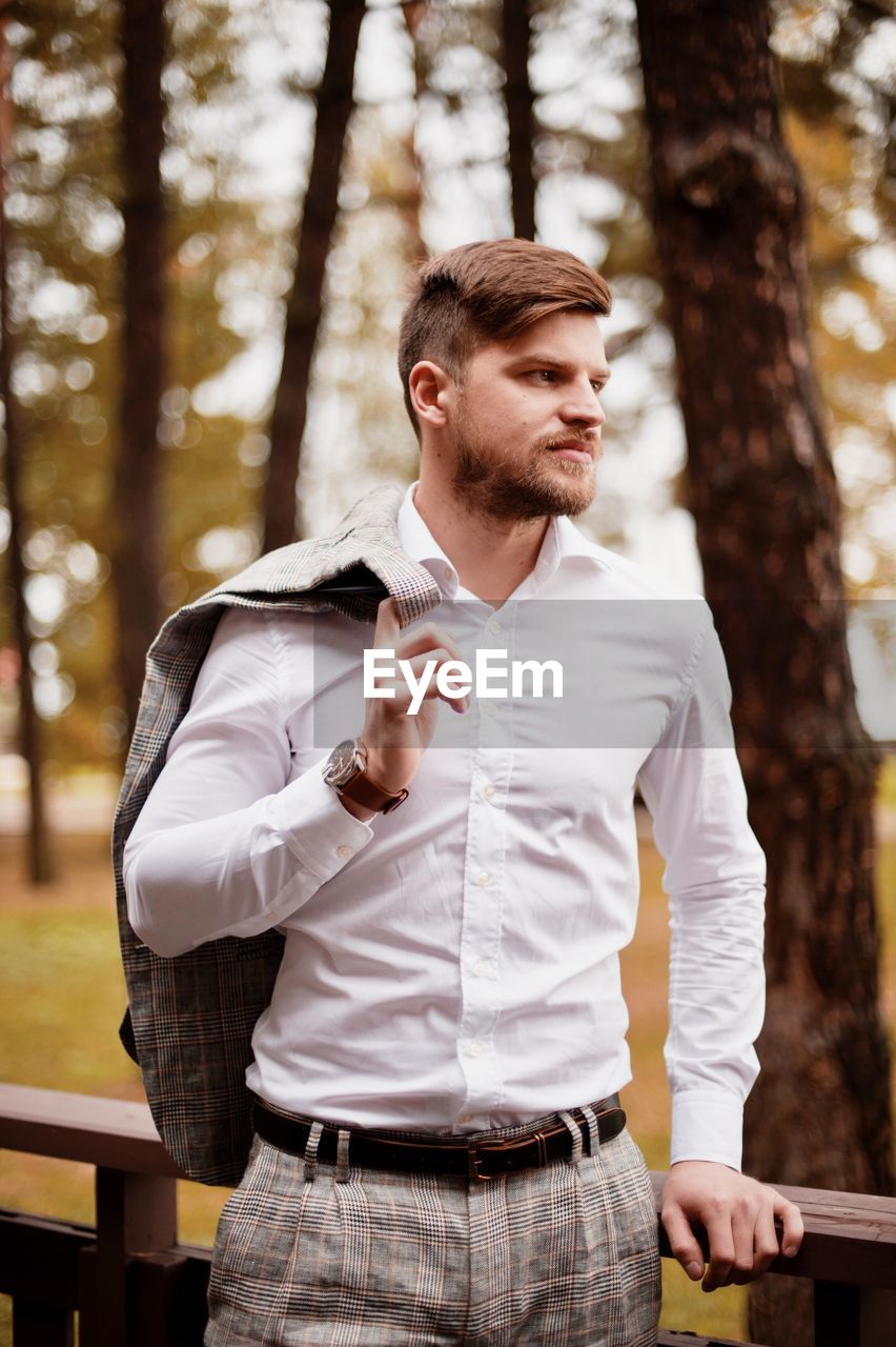 Young man looking away while standing by railing against trees in forest