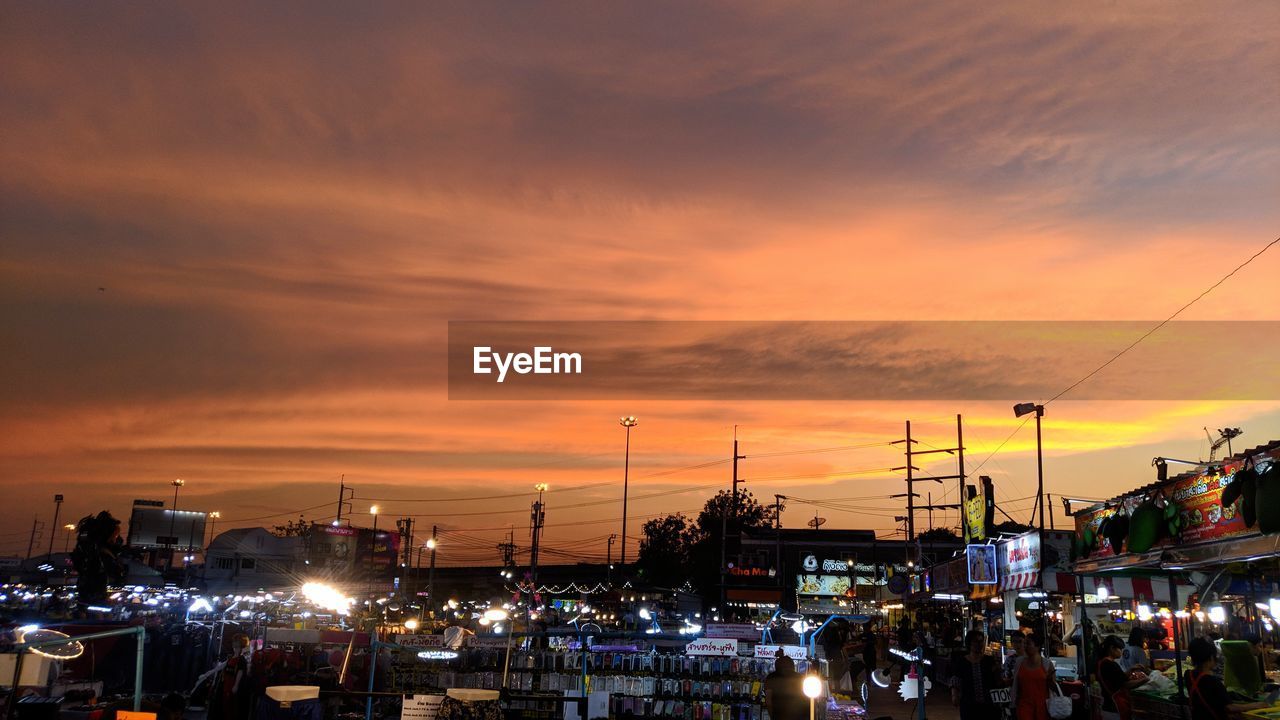 CARS ON ILLUMINATED STREET BY BUILDINGS AGAINST SKY DURING SUNSET