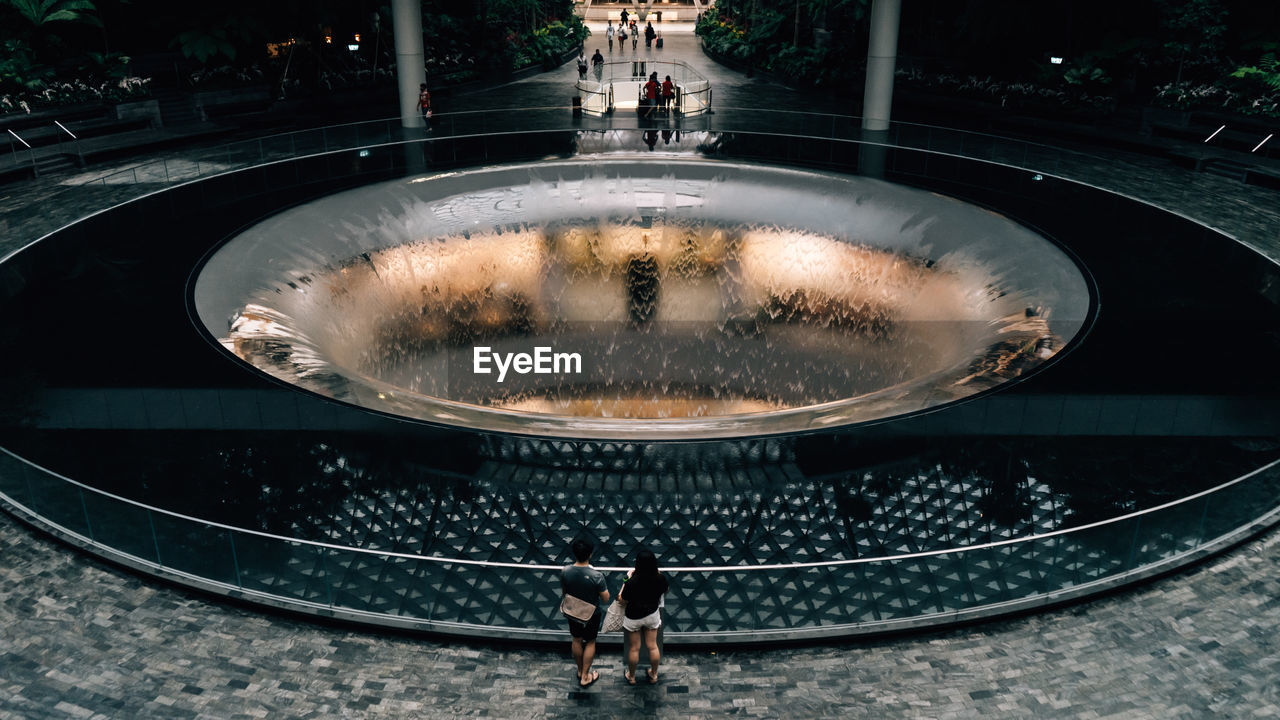 REAR VIEW OF MAN STANDING BY FOUNTAIN