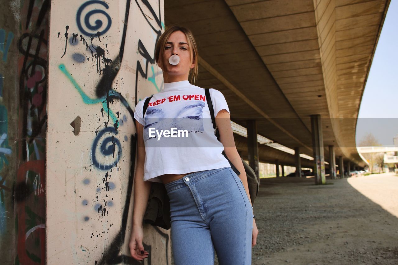 Portrait of young woman blowing bubble gum while standing under bridge