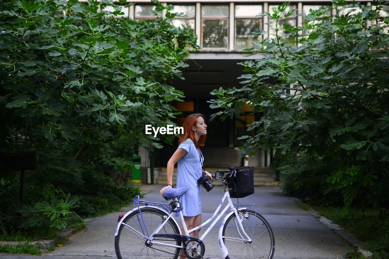 Young woman with bike in front of apartment building and large bush