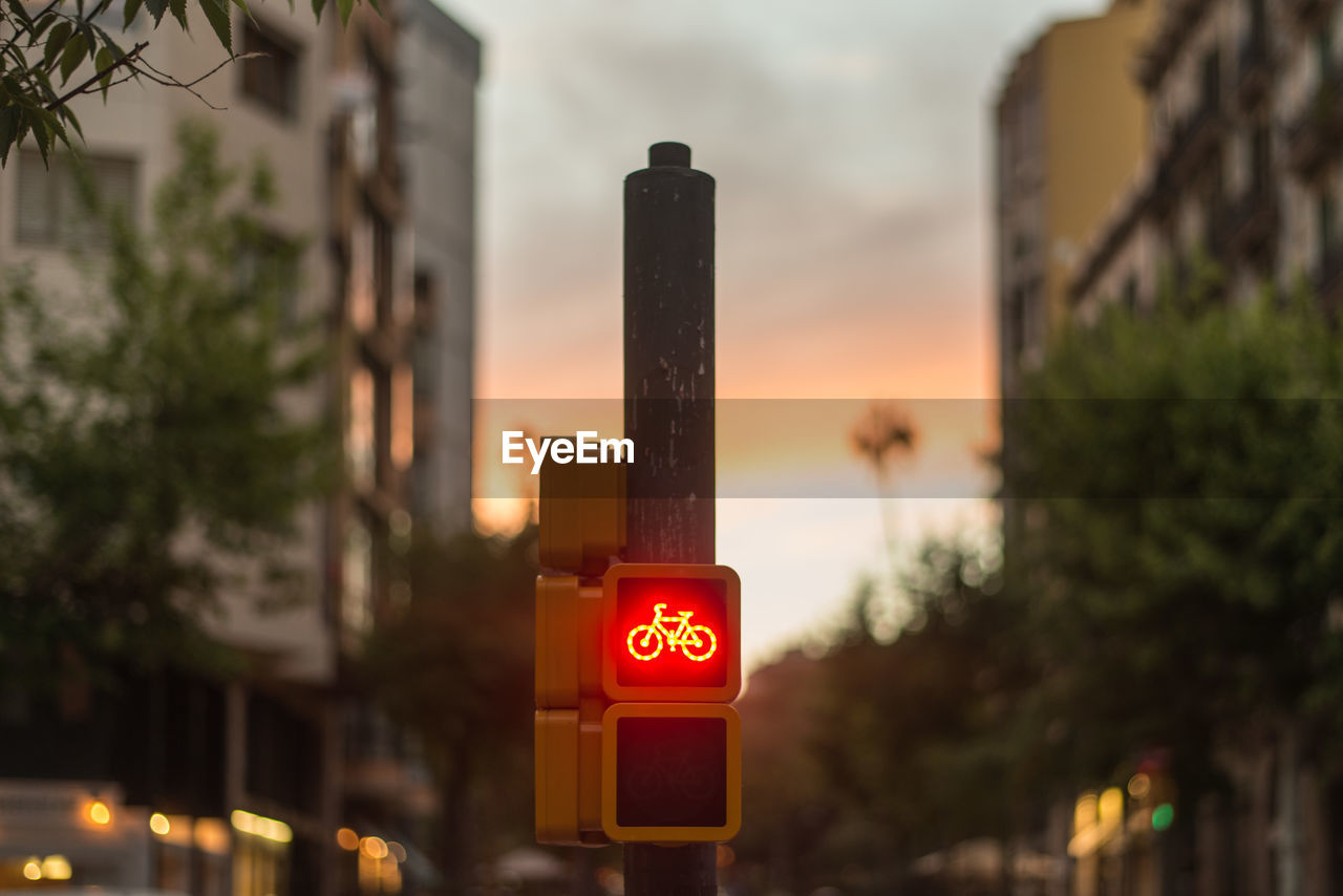 Bicycle sign on pole in city against sky during sunset