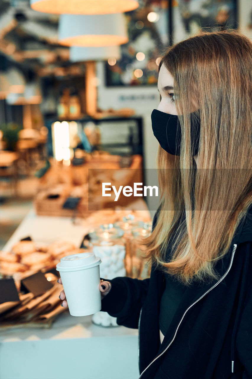 Woman holding coffee cup, standing in queue in a coffee shop. person wearing face mask