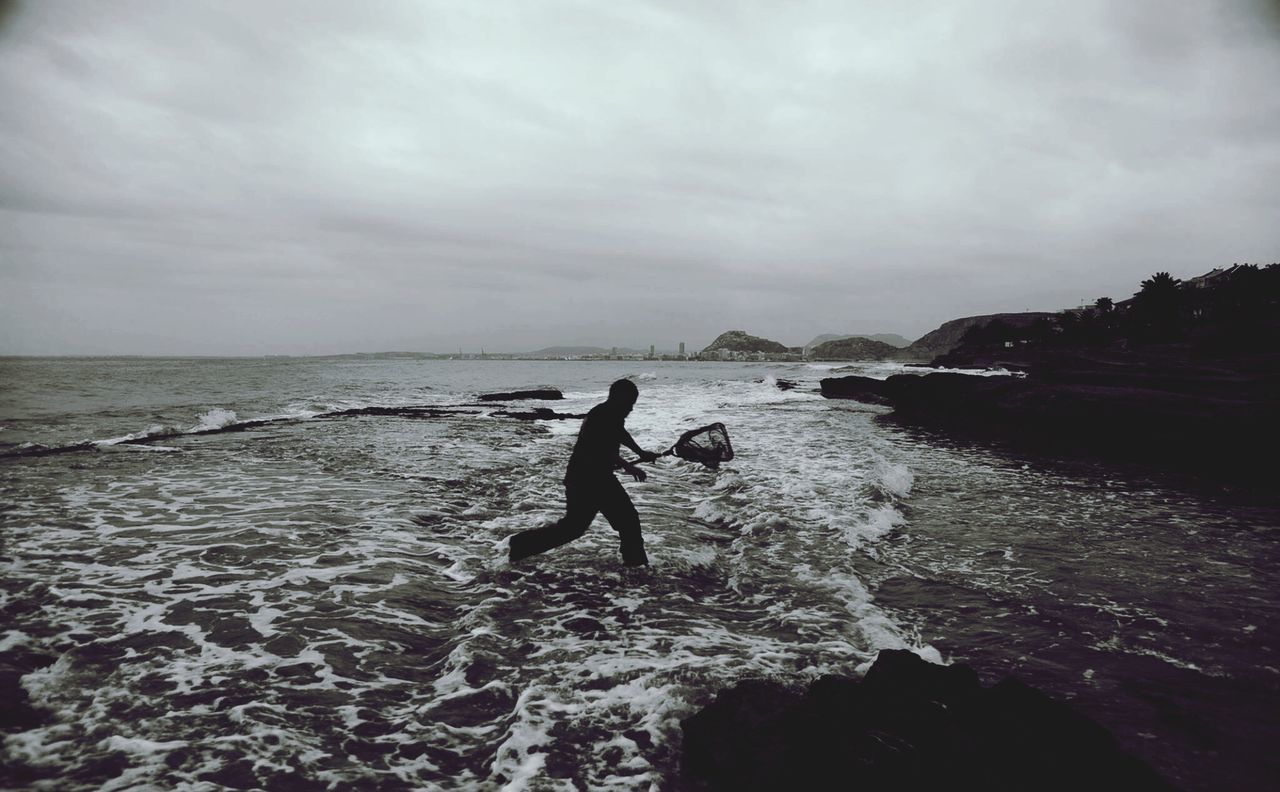 Man fishing with net at sea shore against cloudy sky