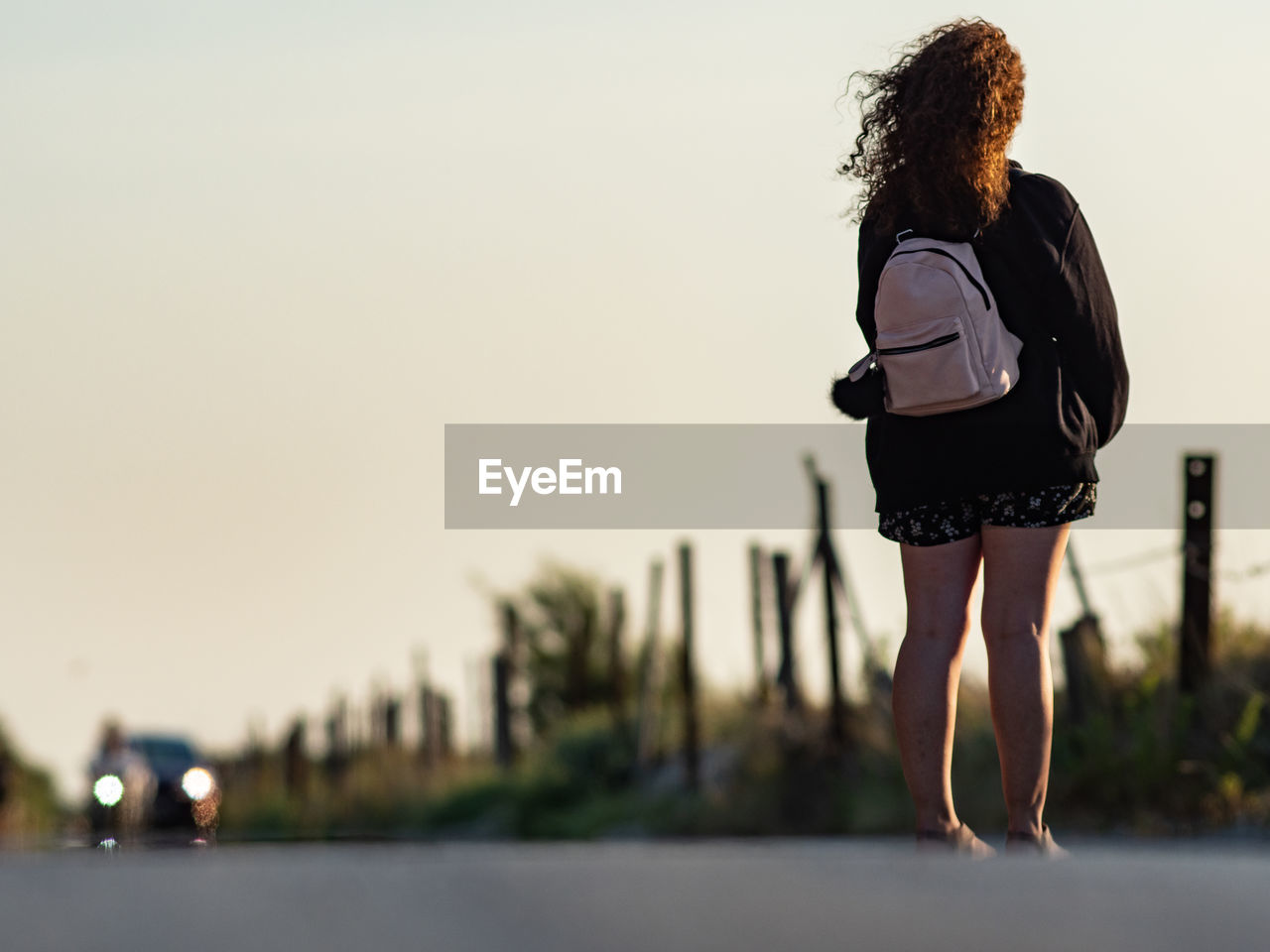 Rear view of woman with backpack standing on road against clear sky at sunset