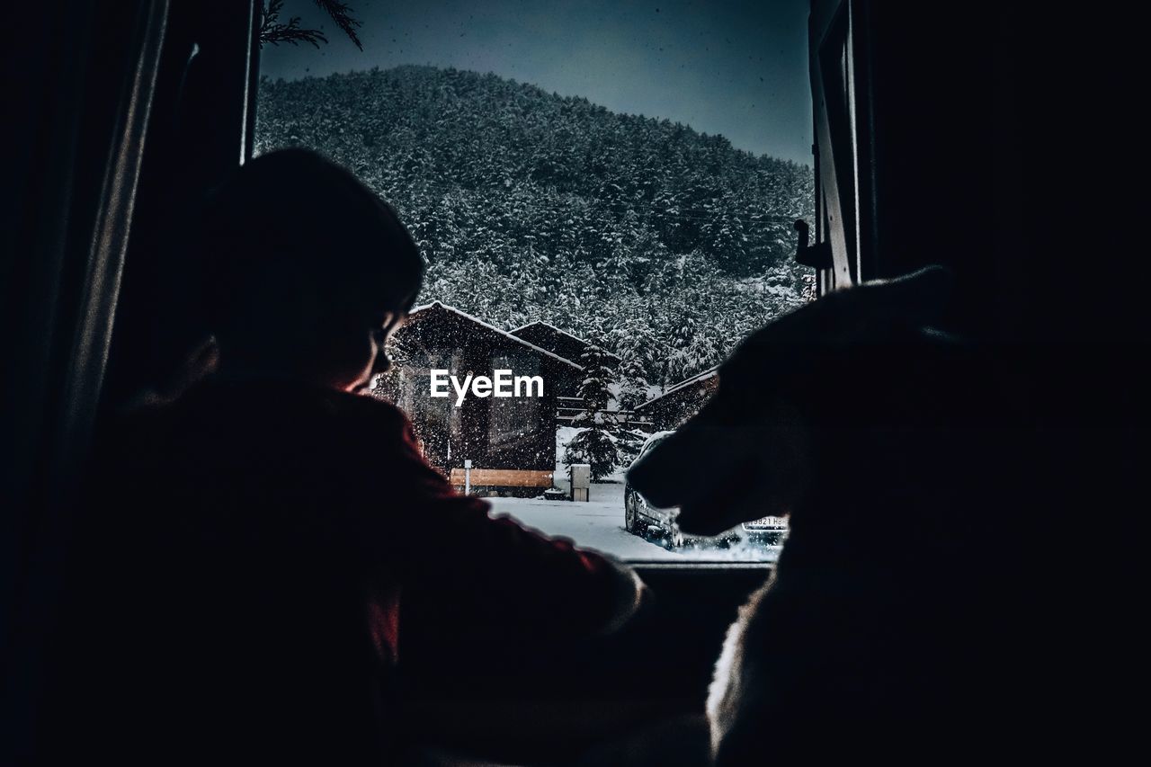 Rear view of boy sitting with dog by window at home