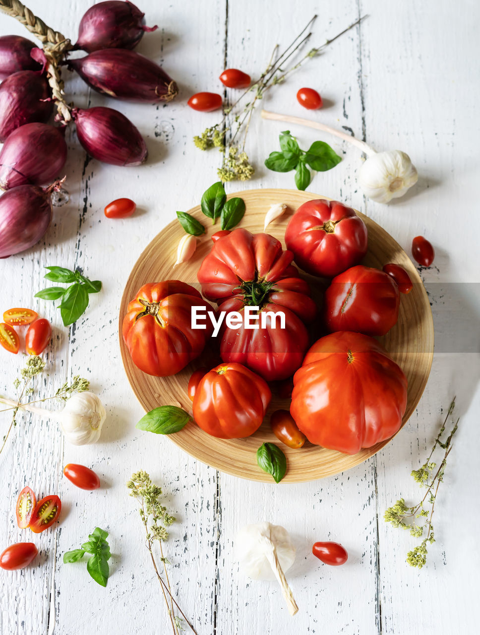 Tomatoes on the plate and white wooden background with oregano, basil leaves, garlic and onion