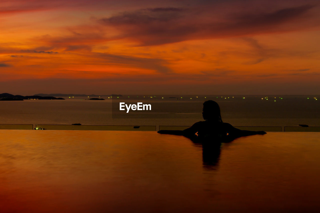 Silhouette woman swimming in infinity pool by sea against sky during sunset