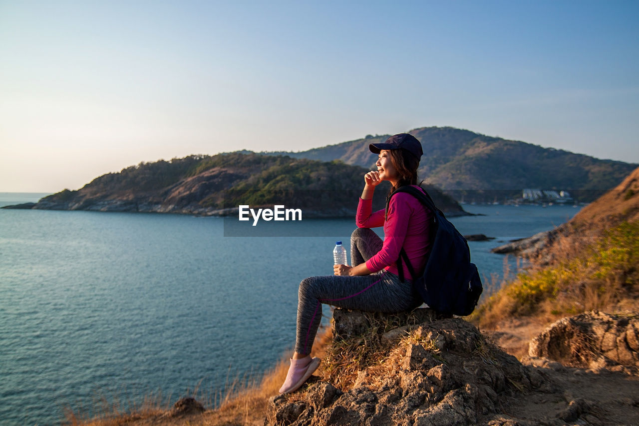 Rear view of woman sitting on rock by sea against sky