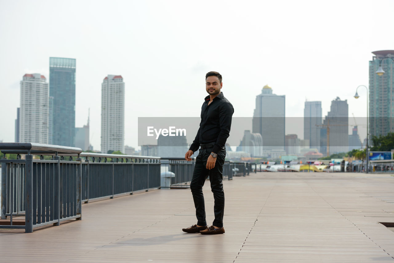 FULL LENGTH OF MAN STANDING ON RAILING AGAINST BUILDINGS IN CITY